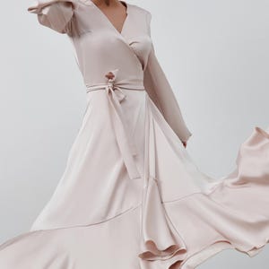 May include: A pale pink wrap dress with long sleeves and a V-neck. The dress has a tie at the waist and a flowing skirt with ruffled edges. The satin fabric gives it an elegant appearance.