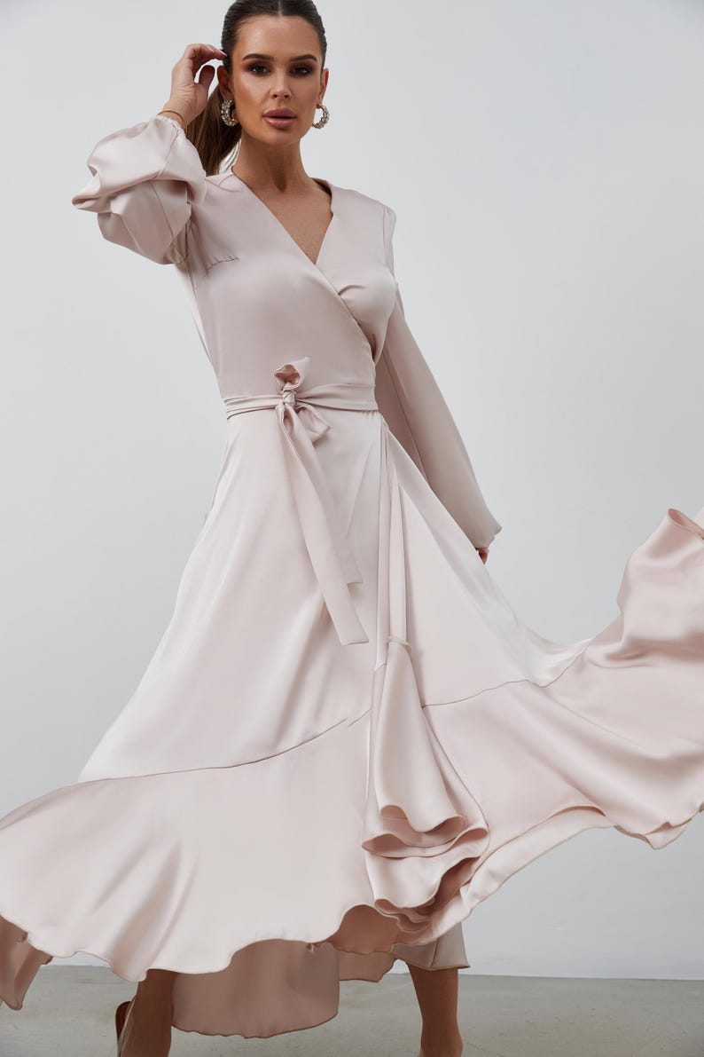 May include: A pale pink wrap dress with long sleeves and a V-neck. The dress has a tie at the waist and a flowing skirt with ruffled edges. The satin fabric gives it an elegant appearance.