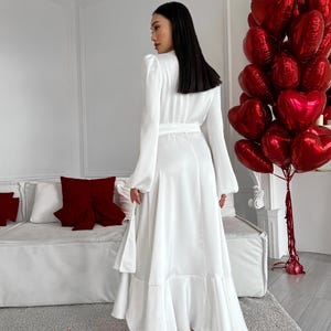 May include: A white satin maxi dress with a flowing skirt and long sleeves. The dress has a belted waist and a ruffled hem. The model is wearing white heels. A bunch of red heart-shaped balloons is in the background.