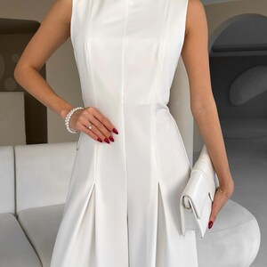 May include: A sleeveless, white dress with a high neckline and pleated skirt. The dress is paired with a matching white clutch. The person is wearing a pearl bracelet and a ring. The nails are painted red.