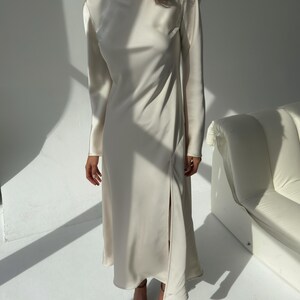 May include: A long, ivory-colored dress with long sleeves and a high side slit. The dress has a simple, elegant design and is made of a smooth, flowing fabric. The dress is paired with brown pointed-toe heels. The overall style is minimalist and chic.