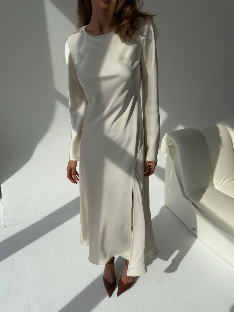 May include: A long, ivory-colored dress with long sleeves and a high side slit. The dress has a simple, elegant design and is made of a smooth, flowing fabric. The dress is paired with brown pointed-toe heels. The overall style is minimalist and chic.