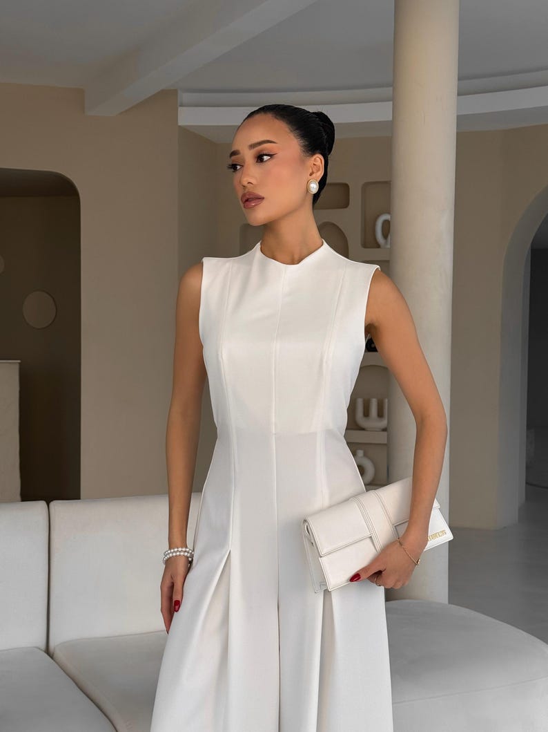 May include: A sleeveless, white jumpsuit with a high neckline and pleated skirt. The outfit is paired with a white clutch and pearl earrings. The model is wearing a pearl bracelet and has red nail polish.