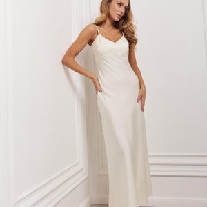 May include: A cream-coloured slip dress with a V-neck and spaghetti straps. The dress is floor-length and paired with red heels. The model is leaning against a white wall.