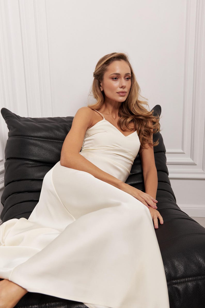 May include: A cream-coloured, floor-length dress with spaghetti straps. The dress has a V-neckline and a flowing skirt. The person is seated on a black leather chair. The background is a white wall with decorative moulding.