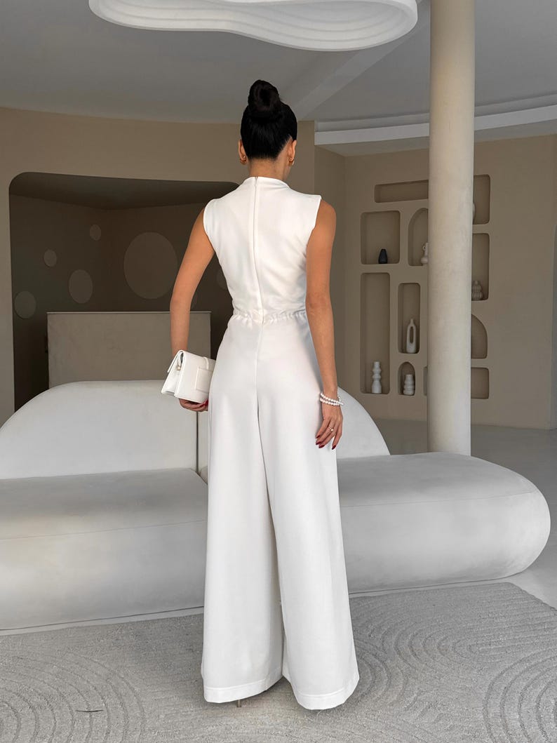 May include: A sleeveless white jumpsuit with wide legs. The outfit features a high neckline and a cinched waist. A white clutch is held in the left hand. The person is facing away from the camera.