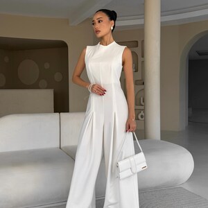 May include: A sleeveless, white jumpsuit with wide-leg pants. The outfit features a high neckline and a matching white handbag. The model is wearing gold sandals and pearl earrings and bracelet.