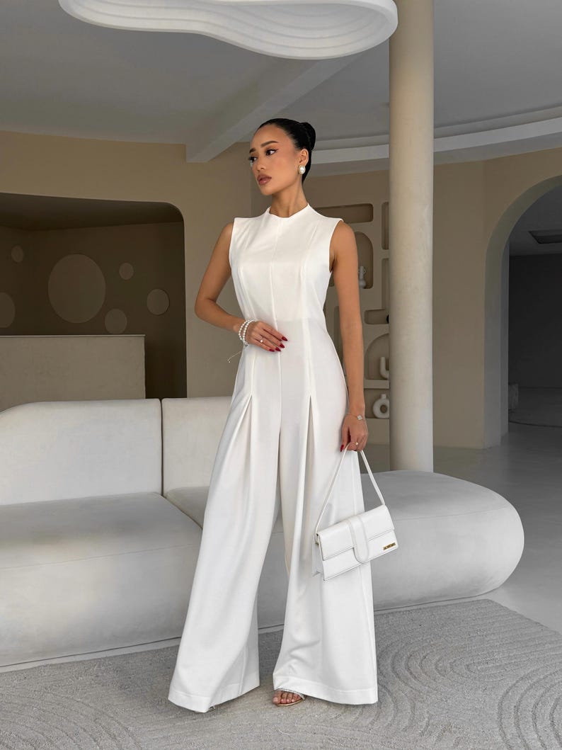 May include: A sleeveless, white jumpsuit with wide-leg pants. The outfit features a high neckline and a matching white handbag. The model is wearing gold sandals and pearl earrings and bracelet.