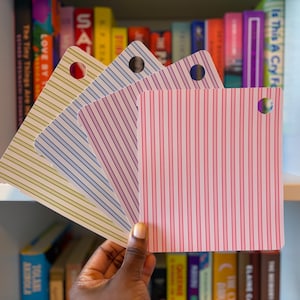 Puede incluir: Un conjunto de cuatro tarjetas cuadradas dispuestas en abanico, cada una con un patrón de rayas verticales distinto en verde lima, azul claro, lavanda y rosa. Cada tarjeta tiene un orificio circular perforado cerca de la esquina superior.