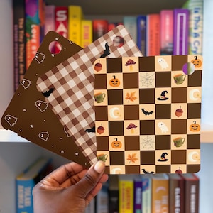 May include: Three square plastic sheets with rounded corners and a hole in the top corner. One sheet is dark brown with white ghost outlines, one is a brown and white gingham check with bats, and one is a brown and cream checkerboard with icons.