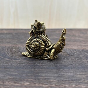 May include: A brass-colored figurine of a snail with a house on its back. The snail has a detailed shell and a smiling face. The house has intricate architectural details. The figurine is placed on a wooden surface.