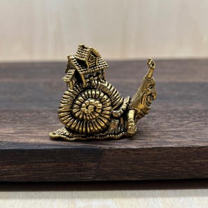 May include: A small, metallic gold-toned figurine shaped like a snail. The snail's shell is replaced by a detailed miniature house with a roof and windows. The snail's head features a distinct, expressive face with a long, curved snout.