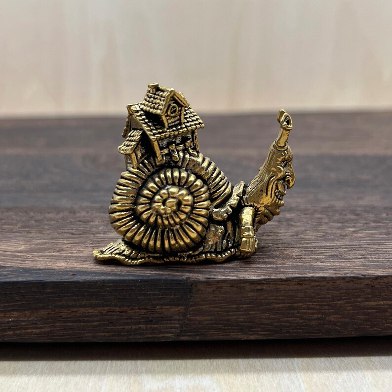 May include: A small, metallic gold-toned figurine shaped like a snail. The snail's shell is replaced by a detailed miniature house with a roof and windows. The snail's head features a distinct, expressive face with a long, curved snout.