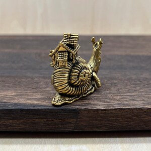 May include: A detailed brass figurine of a snail carrying a miniature house on its back. The snail's shell features intricate spiral detailing, and the house has a chimney and multiple windows. The figurine is set against a dark wood background.