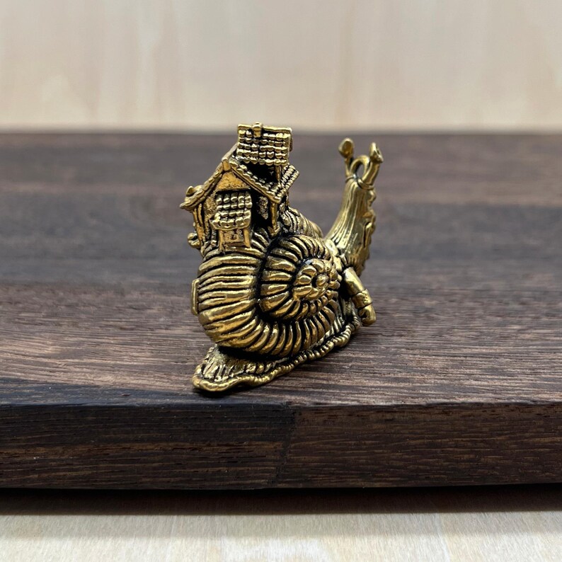 May include: A detailed brass figurine of a snail carrying a miniature house on its back. The snail's shell features intricate spiral detailing, and the house has a chimney and multiple windows. The figurine is set against a dark wood background.