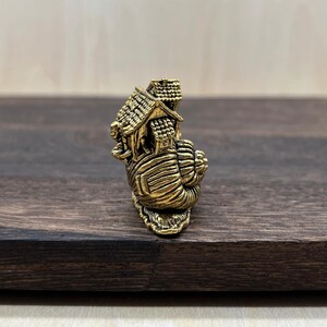 May include: A small, metallic gold-toned figurine shaped like a snail with a house structure resting on its shell. The snail has a textured, spiraled shell base, and the house features a peaked roof with detailed shingle patterns.