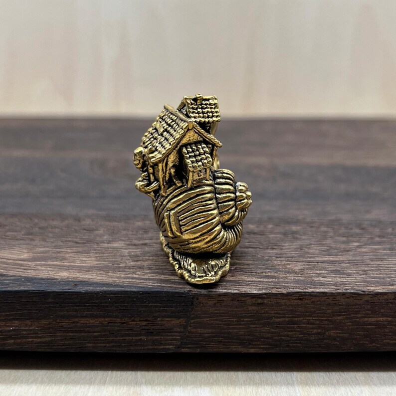 May include: A small, metallic gold-toned figurine shaped like a snail with a house structure resting on its shell. The snail has a textured, spiraled shell base, and the house features a peaked roof with detailed shingle patterns.