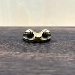 Könnte beinhalten: Eine kleine Froschfigur aus Messing mit detailliertem Design. Der Frosch hat große, runde Augen und einen lächelnden Mund. Die Figur steht auf einer dunklen Holzoberfläche.
