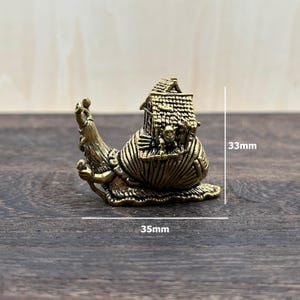 May include: A brass-colored metal figurine of a snail with a house on its shell. The snail has a humanoid face and arms, and the house features a textured roof. The figurine measures 33mm in height and 35mm in width as indicated by the text.
