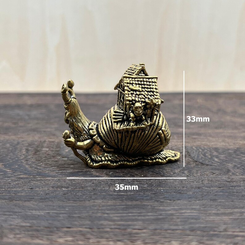 May include: A brass-colored metal figurine of a snail with a house on its shell. The snail has a humanoid face and arms, and the house features a textured roof. The figurine measures 33mm in height and 35mm in width as indicated by the text.