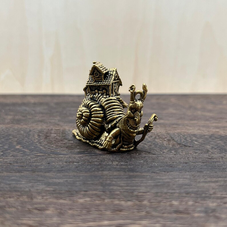 May include: A detailed brass figurine of a snail carrying a house on its back. The snail has intricate detailing, including a textured shell and a smiling face. The house features a detailed roof and windows. The figurine is set against a dark wood background.