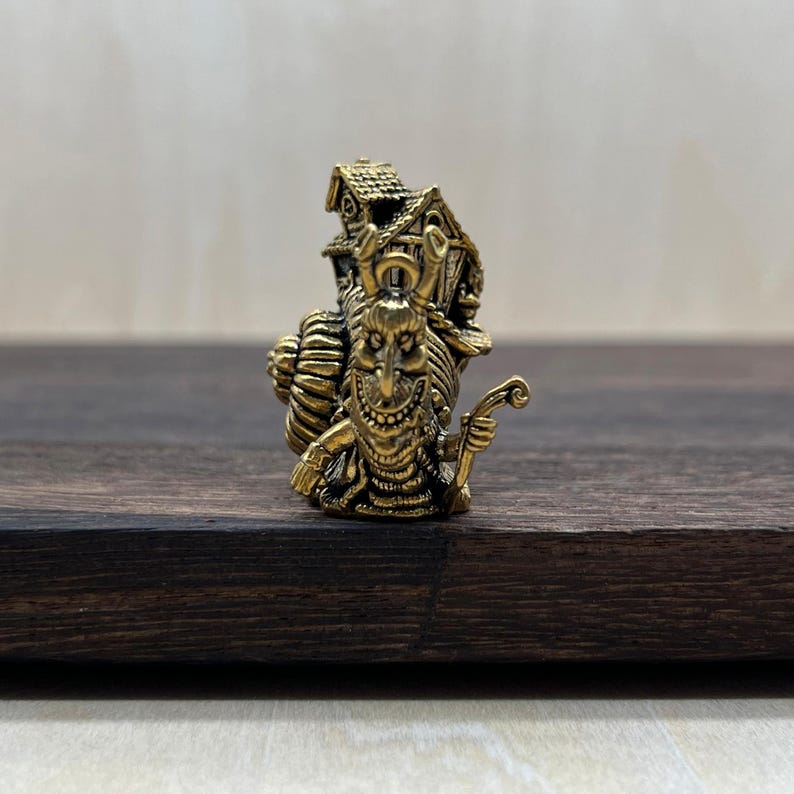 May include: A detailed brass figurine of a whimsical character with a long nose, carrying a house on its back. The figure has a smiling expression and holds a curved staff. The overall design is intricate and detailed.