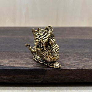 May include: A detailed brass figurine of a snail carrying a house on its back. The house has a textured roof and intricate details. The snail's shell is also textured, and the figurine has a golden hue.