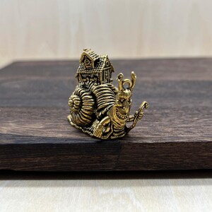 May include: A detailed brass figurine of a snail carrying a house on its back. The snail has a smiling face and holds a small staff. The house has intricate details, including a roof and windows. The figurine is set on a dark wooden surface.