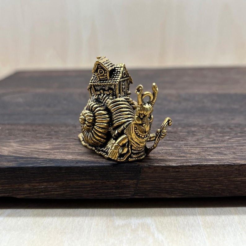 May include: A detailed brass figurine of a snail carrying a house on its back. The snail has a smiling face and holds a small staff. The house has intricate details, including a roof and windows. The figurine is set on a dark wooden surface.