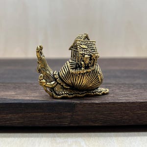 May include: A detailed brass figurine of a snail carrying a house on its back. The snail has intricate detailing, with a textured shell and a whimsical house featuring a roof and small figures. The figurine is set on a dark wooden surface.