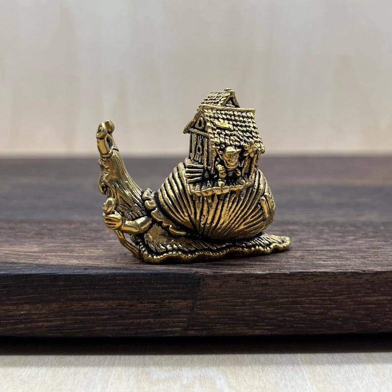 May include: A detailed brass figurine of a snail carrying a house on its back. The snail has intricate detailing, with a textured shell and a whimsical house featuring a roof and small figures. The figurine is set on a dark wooden surface.