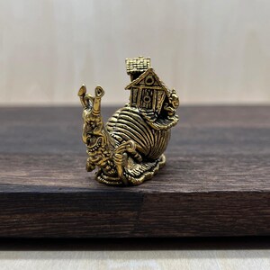 May include: A small, metallic gold-toned figurine of a snail with a detailed, textured shell that resembles a miniature house with a roof and walls. The snail has a prominent face with two antennae and a curved, expressive body structure.