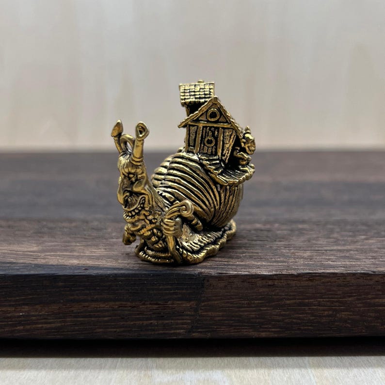 May include: A small, metallic gold-toned figurine of a snail with a detailed, textured shell that resembles a miniature house with a roof and walls. The snail has a prominent face with two antennae and a curved, expressive body structure.