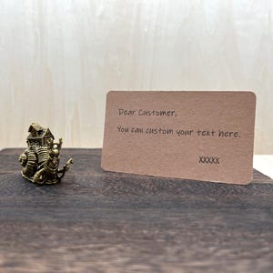 May include: A bronze-colored snail figurine with a house on its back, next to a brown card with the text "Dear Customer, You can custom your text here." The snail is on a dark wooden surface.