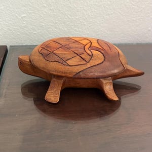 May include: A wooden turtle figurine with a detailed shell design. The turtle is carved from a light brown wood, with darker brown accents on the shell. The figurine has four legs and a small head, and is sitting on a dark surface.