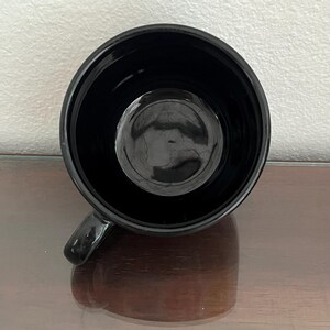 May include: A glossy black mug with a rounded interior and a small handle. The mug's interior reflects the light, creating a circular highlight. The mug sits on a reflective surface.