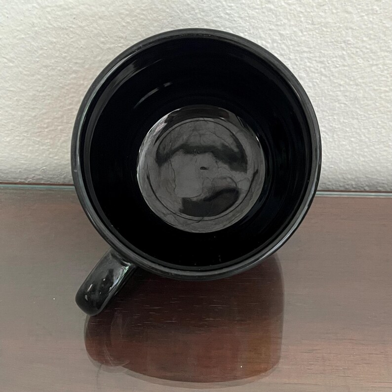 May include: A glossy black mug with a rounded interior and a small handle. The mug's interior reflects the light, creating a circular highlight. The mug sits on a reflective surface.