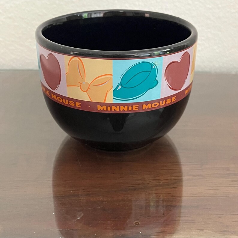 May include: Black ceramic bowl with a colorful band featuring Minnie Mouse-themed graphics. The band includes hearts, bows, and a hat, with the words "MINNIE MOUSE" in red. The bowl has a glossy finish and a dark interior.