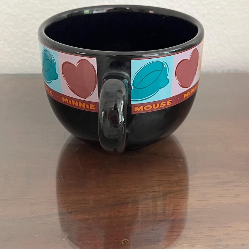 May include: Black ceramic Minnie Mouse mug with a rounded shape and a large handle. The mug features a decorative band with hearts, teal shapes, and the words "Minnie Mouse" in red. The interior is dark blue.
