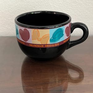 May include: Black ceramic Minnie Mouse mug with a rounded shape and a handle. The mug features a colorful band with Minnie Mouse-themed graphics, including hearts, bows, and hats. The words "Minnie Mouse" are printed on the band.