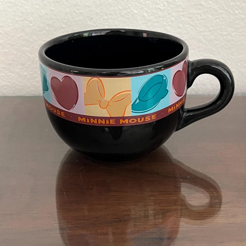 May include: Black ceramic Minnie Mouse mug with a rounded shape and a handle. The mug features a colorful band with Minnie Mouse-themed graphics, including hearts, bows, and hats. The words "Minnie Mouse" are printed on the band.