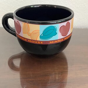 May include: Black ceramic Minnie Mouse coffee cup with a black handle. The cup features a colorful band with Minnie Mouse-themed graphics, including hearts, bows, and a hat. The words "Minnie Mouse" are printed around the band.