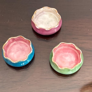 May include: Three small, decorative ceramic bowls in shades of pink, blue, and green. Each bowl has a scalloped rim with a gold accent and a contrasting interior color. The bowls are arranged on a dark brown surface.