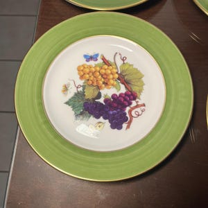 May include: A round ceramic plate with a wide lime green rim and a gold-toned edge. The center features a white background decorated with a cluster of yellow and purple grapes, green vine leaves, and two small butterflies in blue and yellow.