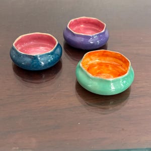 May include: Three small, handmade ceramic bowls in shades of teal, purple, and green. Each bowl has a contrasting interior color of pink or orange. The bowls are round with a smooth, glossy finish, perfect for holding small items or as decorative accents.