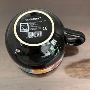 May include: Black cappuccino mug with a handle, featuring a colorful design around the base. The bottom of the mug displays the "applause" logo, Disney branding, and product information. Dishwasher and microwave safe.