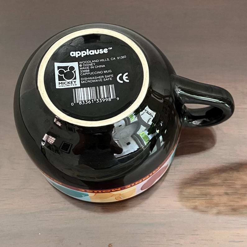May include: Black cappuccino mug with a handle, featuring a colorful design around the base. The bottom of the mug displays the "applause" logo, Disney branding, and product information. Dishwasher and microwave safe.