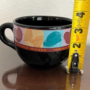 May include: Black ceramic Minnie Mouse teacup with a handle. The cup features a colorful band with hearts, a bow, and a hat design. The words "Minnie Mouse" are printed on the cup.