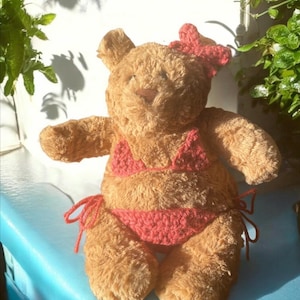 May include: A plush brown teddy bear wearing a red crocheted bikini and a matching bow. The bear is positioned against a light blue surface, with sunlight casting shadows from nearby plants. The bear's fur is soft and textured.