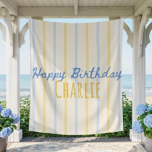 May include: A striped banner with the text "Happy Birthday CHARLIE" in blue and yellow. The banner is hanging on a white porch overlooking the ocean. Blue hydrangea plants in gray pots flank the banner.